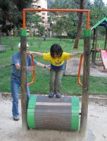 Un enfant joue sur un tonneau en bois qui tourne, aidé par un adulte.
