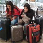 Deux femmes assises sur des sièges, dans une gare avec 4 valises autour d'elles. Elles ont leur téléphone à la main.