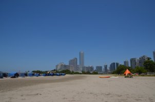 Gratte-ciels au fond, plage de sable devant avec, sur la gauche, des parasols bleus alignés, et sur la droite, 1 parasol pointu orange.