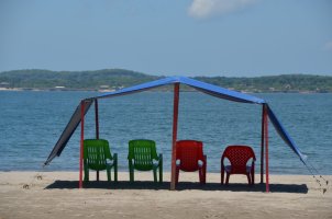 Parasol bleu ouvert abritant 2 chaises vertes et 2 chaises rouges, au bord de la mer.
