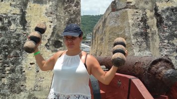 Carole se faisant les muscles avec des boulets en bois devant un canon.